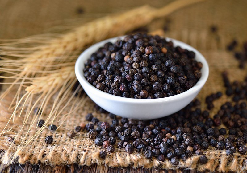 black peppers in a small white bowl on a piece of sack cloth with loose peppercorns around it and a couple of wheat stalks