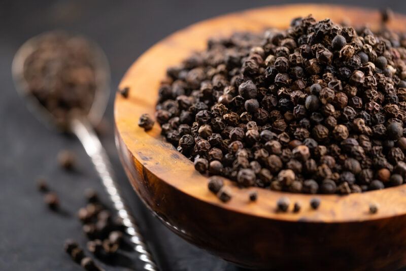 closeup image of a wooden bowl full of black peppercorns with a silver spoon beside it and loose black peppercorns scattered around