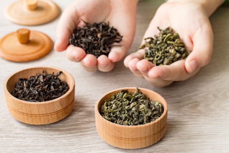 a couple of wooden bowl with loose black and green tea leaves, a couple of hands each holding loose black and green tea leaves