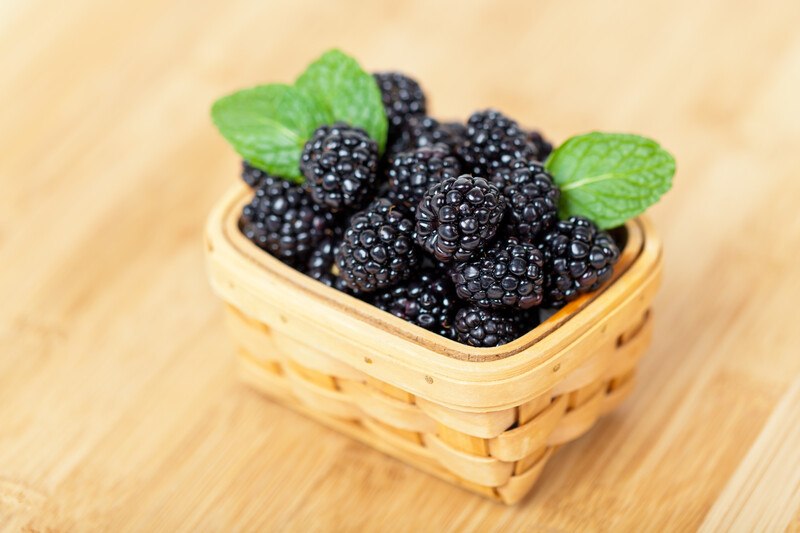 on a light brown wooden surface is a small square woven basket full of blackberries with 3 mint leaves