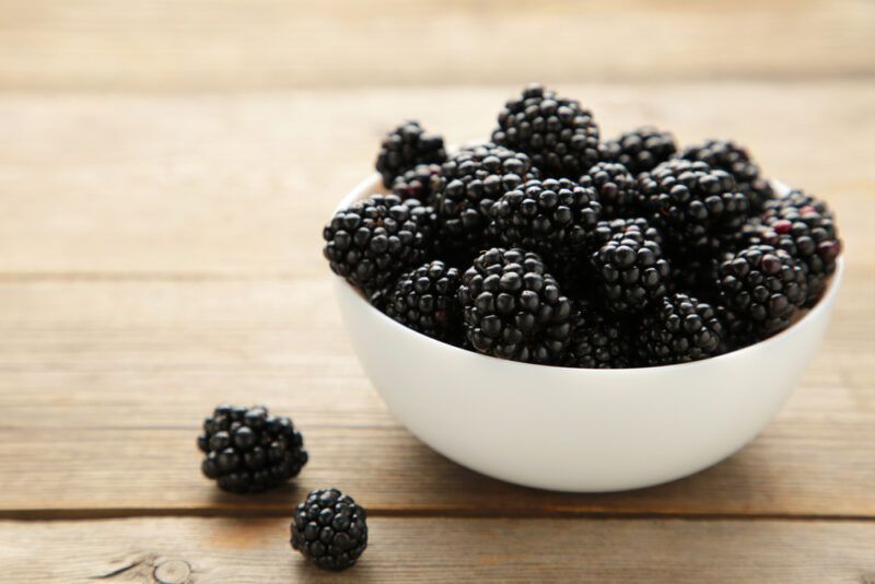 a wooden surface is a white bowl full of blackberries with a couple of loose blackberries beside it