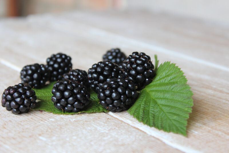 on a light-colored wooden surface are blackberries with a couple of leaves