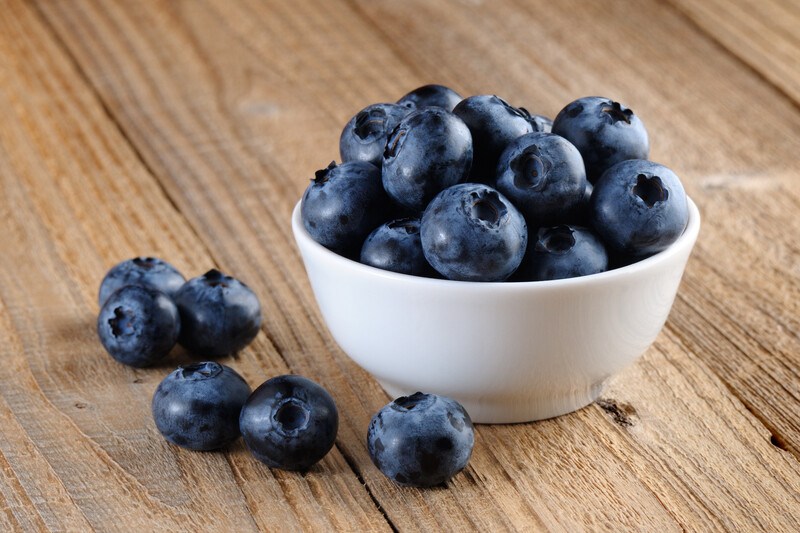 A white dish of blueberries and a few loose blueberries rest on a wooden surface.