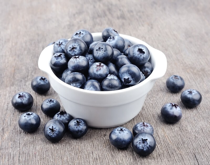 on a dark wooden surface is a ramekin full of blueberries with loose blueberries around it