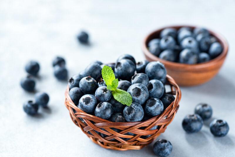 on a white concrete surface is a basket full of blueberries and a clay bowl full of blueberries in the back, with loose blueberries around it