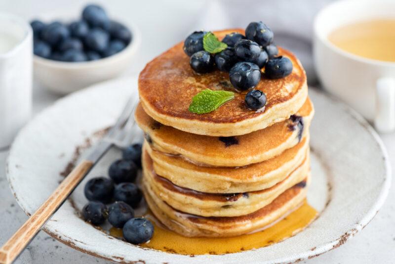 a beautifully plated pile of blueberry almond pancakes with fresh blueberries on top and beside it, with a fork with wooden handle and syrup, a small white bowl of fresh blueberries and a white cup with tea at the back