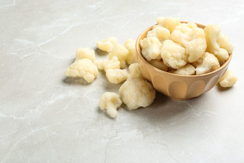 on a concrete surface is a brown ceramic bowl with boiled cauliflower florets, around it are loose boiled cauliflower florets as well