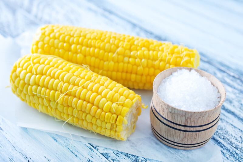 on a rustic wooden surface is a couple of boiled sweet corn on a white parchment paper with a small wooden bowl full of rock salt