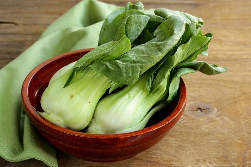 a closeup image of brown wooden bowl with bok choy with a green kitchen towel, resting on a wooden surface