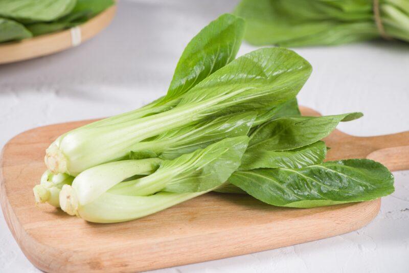 on a white surface is a wooden chopping board with fresh bok choy on it, at the back are partial image of bok choy as well 