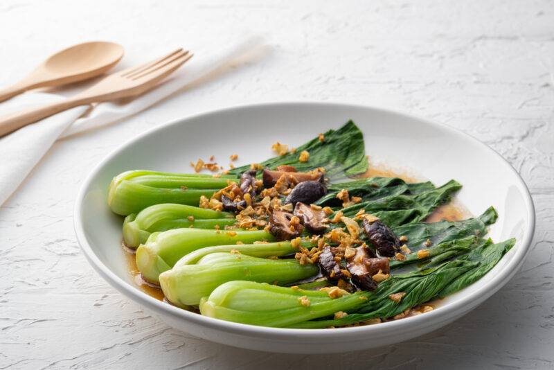 a beautifully plated steamed bok choy with fried garlic and mushroom on top and soy and olive oil sauce, at the back is a spoon and fork resting on top of a white table napkin