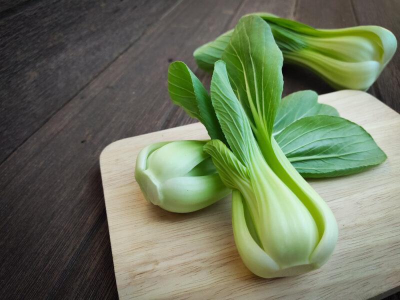 on a dark brown wooden surface is a light brown wooden chopping board with bok choy, a head of bok choy can be seen at the back as well