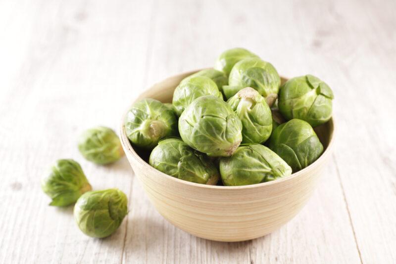 on a white wooden surface is a light brown bowl full of Brussel sprouts, with 3 loose Brussels sprouts beside it