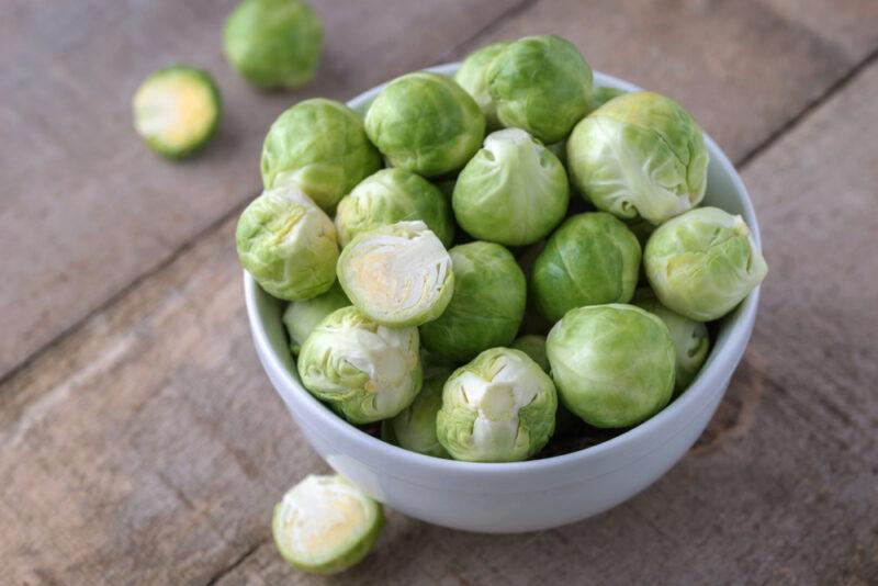 on rustic looking wooden surface is a white bowl full of Brussels sprouts with three loose Brussels sprouts