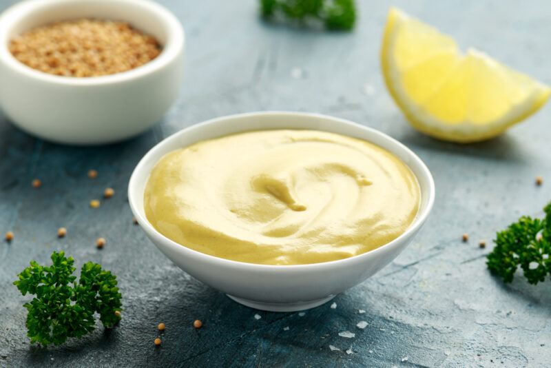 on a dark concrete surface is a white bowl of Dijon mustard with loose parsley, salt, peppercorns, and a lemon wedge around it, at the back is a ramekin full of mustard seeds