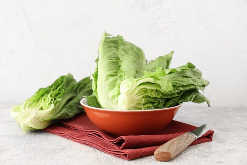 a rust-colored bowl with white rim full of Romaine lettuce, beside it is another Romaine lettuce, resting on a red table napkin wit a knife beside it 