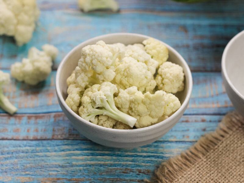 on a rustic-looking blue wooden surface is a white bowl with cauliflower florets with loose cauliflower florets around it