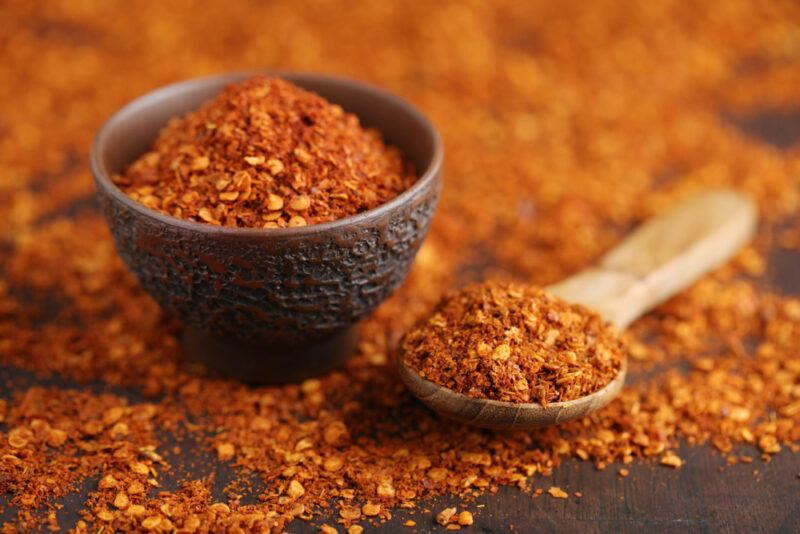 on a dark surface is a spread of cayenne pepper with a small black bowl and wooden spoon on it, both full of cayenne pepper as well