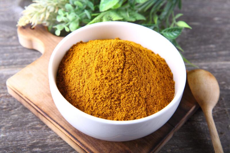 on a wooden surface is a wooden chopping board with white bowl full of curry powder, with a wooden spoon beside it and a sprig of fresh herbs at the back