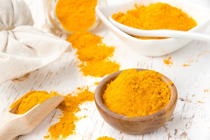 on a white rustic wooden surface is a wooden scoop, wooden bowl, white ceramic bowl, and glass jar, all full of curry powder