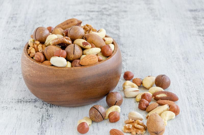 on a whitish wooden surface is a brown wooden bowl full of different kinds of nuts, with loose nuts at the foreground