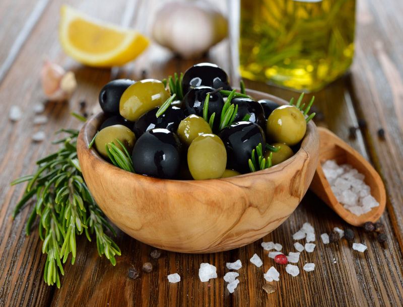 a bowl of olives with sprigs of rosemary on top of a wooden table.