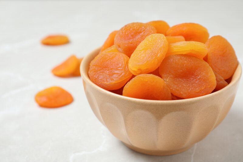 on a white surface is a brownish bowl full of dried apricot with three loose dried apricots at the back