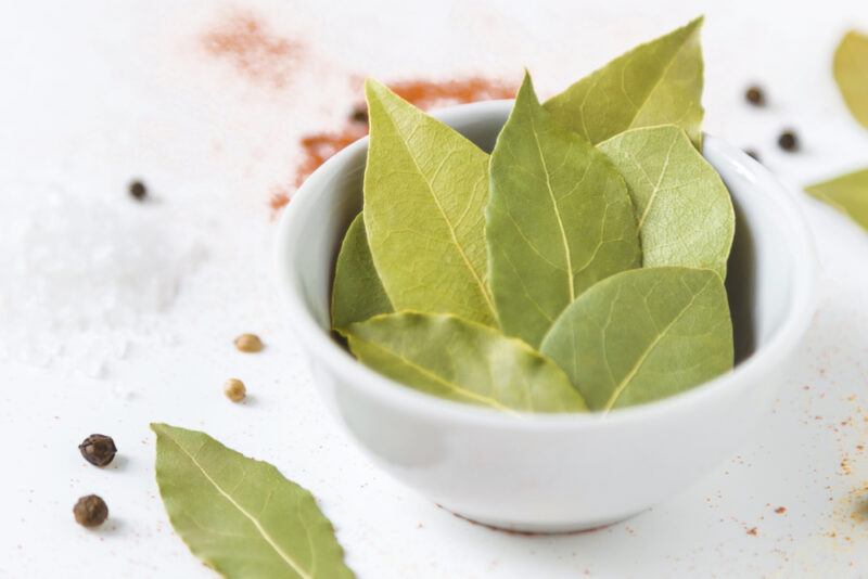 on a marble surface is a white bowl full of dried bay leaves with loose pepper corns and dried bay leaves around it