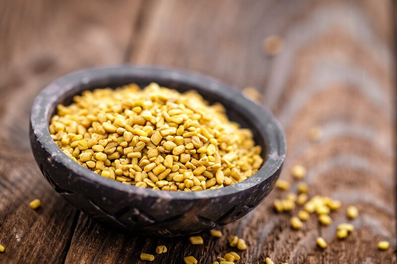 on a rustic wooden surface is a black bowl of fenugreek seeds with loose fenugreek seeds around it