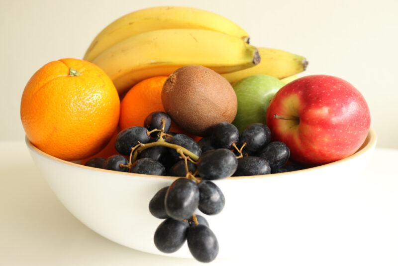 on a white surface is a white bowl of fruits including bananas, orange, grapes, kiwi, and green and red apples