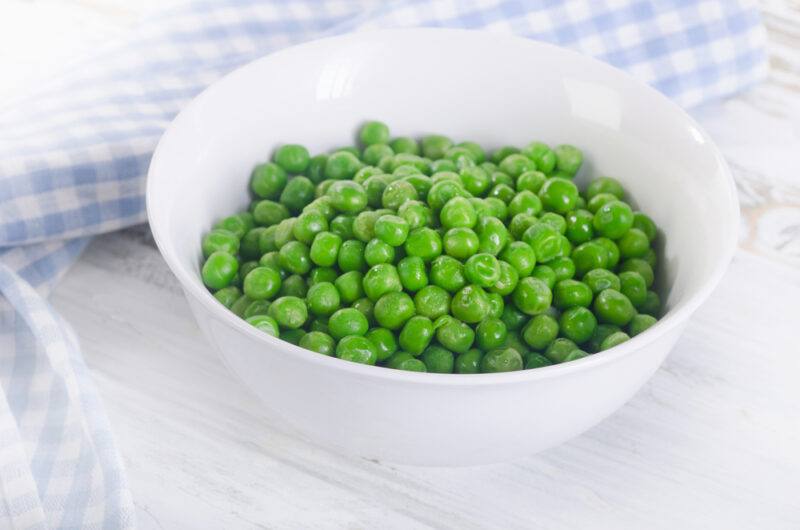 a white surface with a white bowl full of green peas with blue and white table napkin at the back