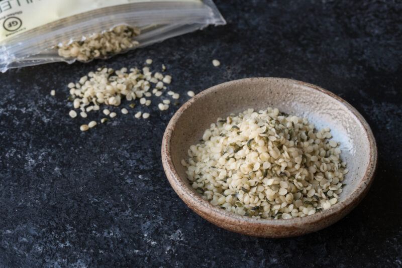 on a dark concrete surface is a brown bowl with hemp hearts, with a bag of hemp hearts with some of it pouring out can be seen partially at the back