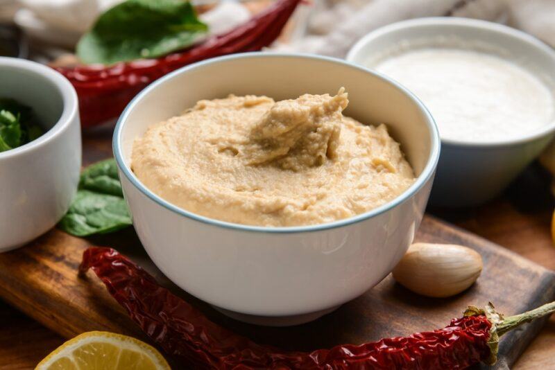 on a white bowl of hummus resting on a wooden chopping board with garlic, chili, a sliced of lemon, and herbs beside it