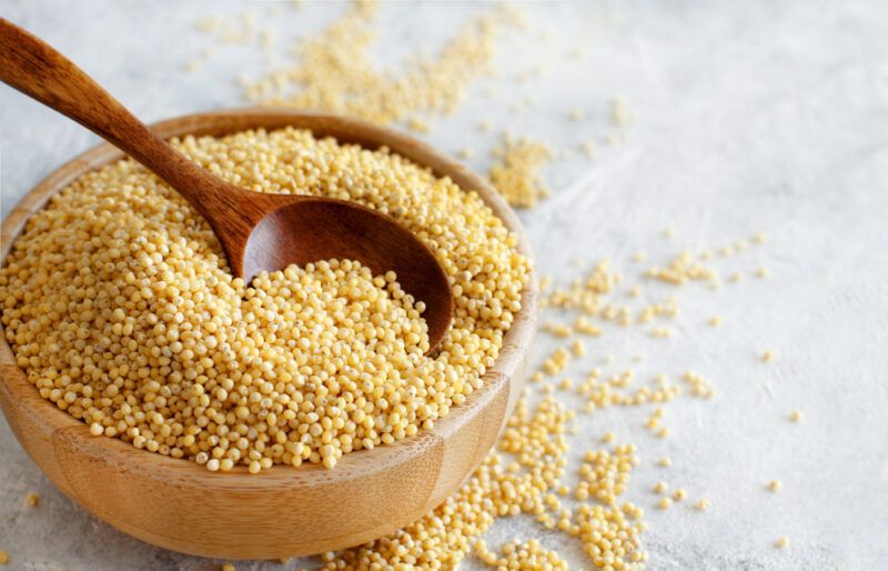 on a marble surface is a brown ceramic bowl of millet with dark brown wooden spoon in it, loose millet grains can be seen around it