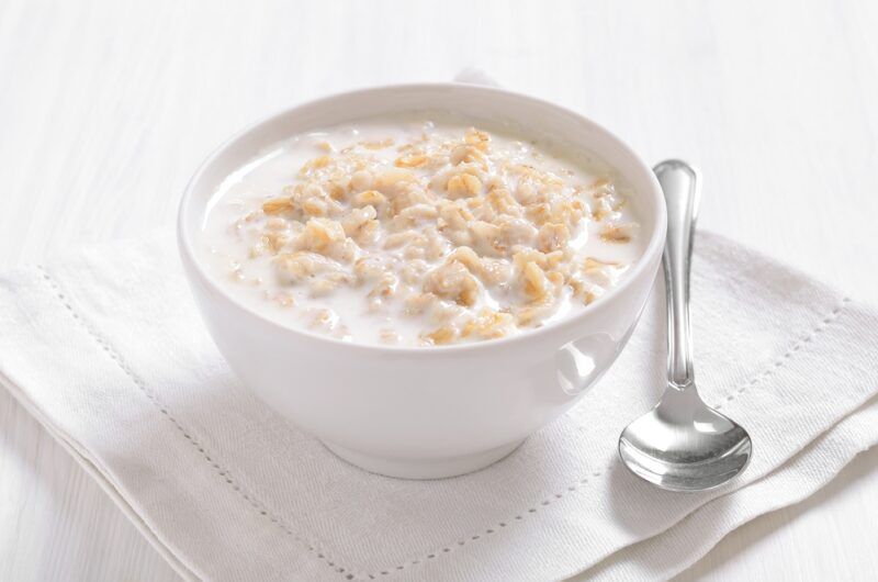 on a white surface is a white bowl of oatmeal with a silver spoon beside it, resting on a white table napkin