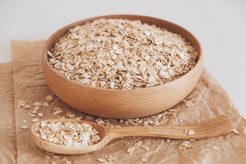 a closeup image of a wooden bowl with oats resting on top of a brown paper with a wooden spoon with oats in front of it, around it are loose oats