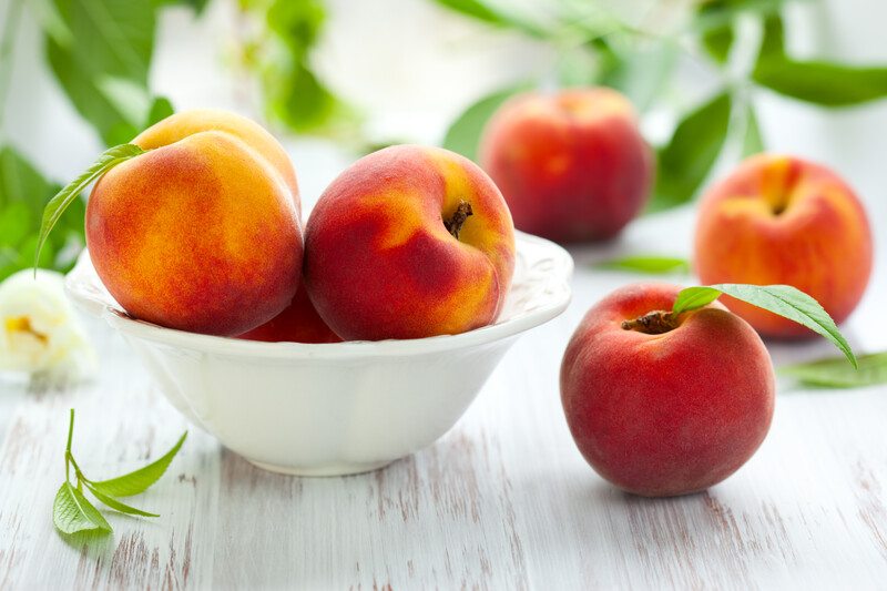 bowl of peaches | Food For Net white wooden surface with a bowl full of fresh whole peaches, with loose peaches with leaves still attached around it