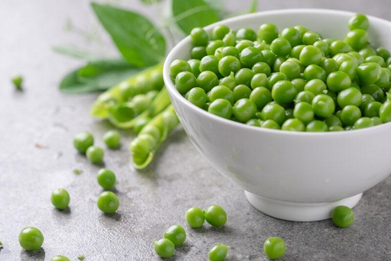 A white bowl of peas with various peas scattered on the table