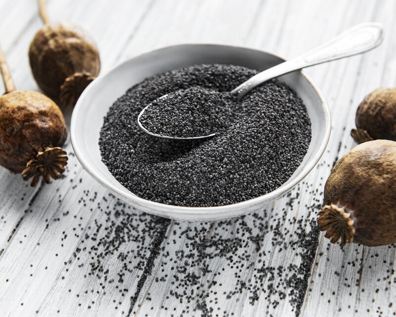 a white rustic wooden surface with a white bowl of poppy seeds, with loose poppy seeds and whole poppy pods around it