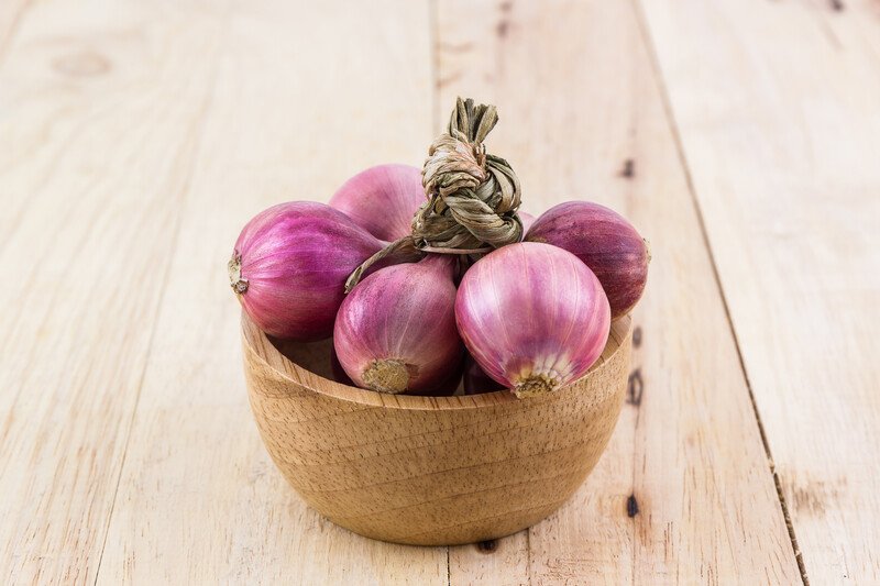 on a light brown wooden surface is a wooden bowl full of whole fresh red onions