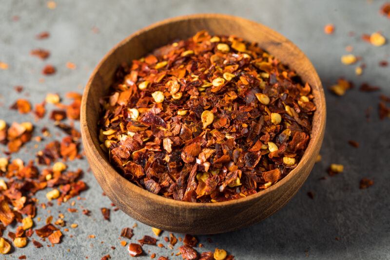 a wooden bowl with red pepper flakes, with loose red pepper flakes around it