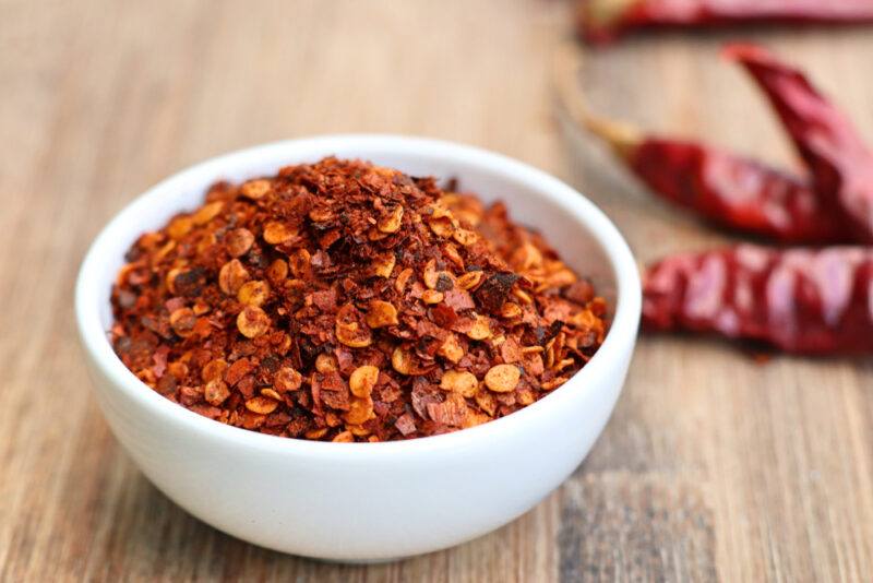 on a wooden surface is a white bowl of red pepper flakes with dried chilis at the back