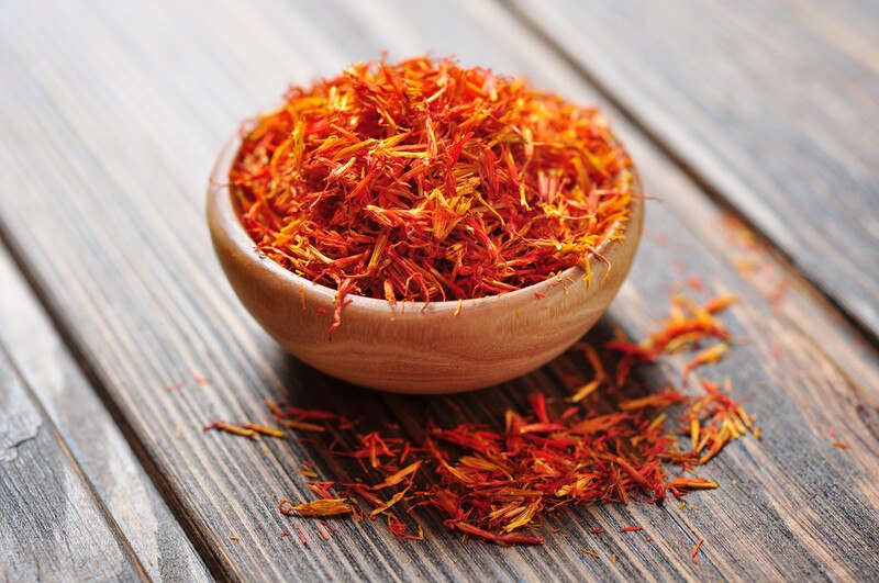 on a rustic wooden surface is a wooden bowl full of saffron threads with loose saffron threads around it