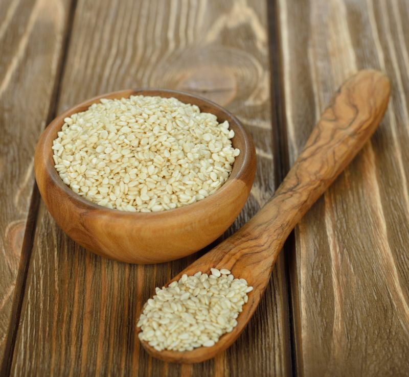 a small wooden bowl filled with white sesame seeds, a wooden spoon with sesame seeds on wooden table.