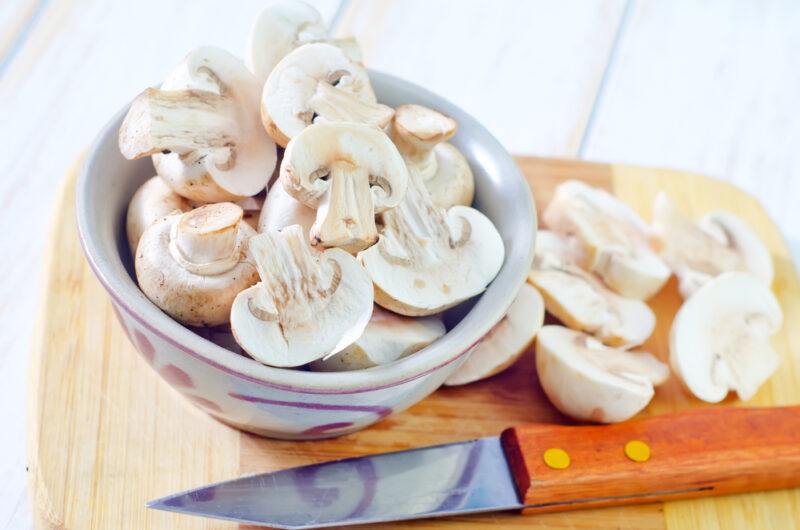 on a white wooden surface is a brown chopping board with a bowl of sliced mushroom, with a knife and loose mushroom slices beside it