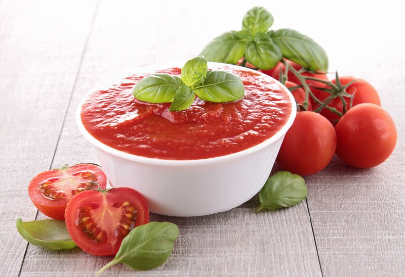 on a white wooden surface is a white ceramic bowl full of tomato sauce with herbs on top, with sliced and whole fresh tomatoes around it