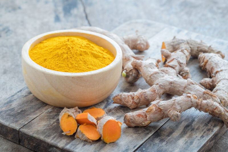 on a wooden board is a white bowl of turmeric powder with fresh turmeric roots around it