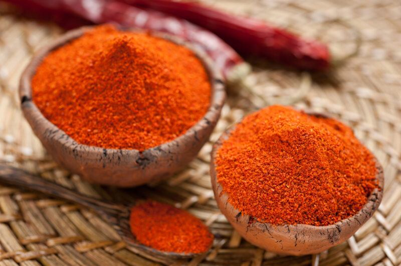 on a native weave platter is a couple of clay bowls full of chili powder with wooden ladle beside it full of chili powder as well, and a couple of dried chili at the back