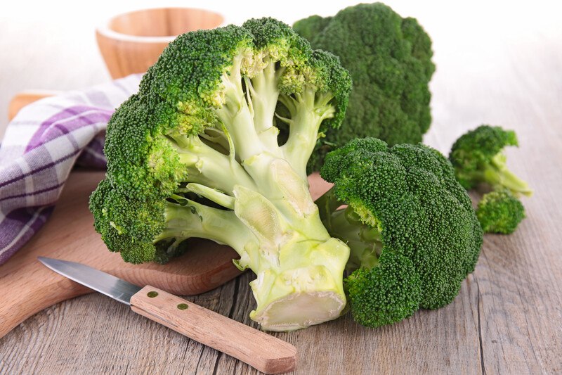 on a wooden surface is three broccoli heads with one which stalk or stem is showing, beside it is a knife, a wooden chopping board, white and lavender table napkin and wooden bowl at the back