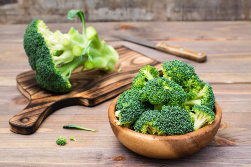 on a wooden surface is a wooden bowl full of broccoli florets, at the back is a wooden chopping board with a broccoli head on it and a knife beside it